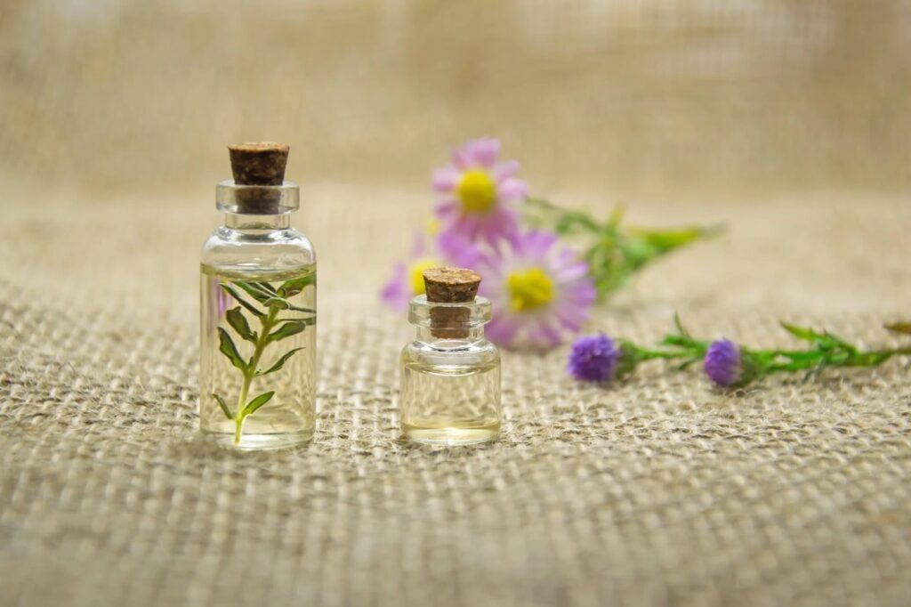 A imagem apresenta dois frascos de óleos essenciais muito utilizados em aromaterapia, com ramos de flores dentro, e flores de lavanda apresentadas ao fundo.