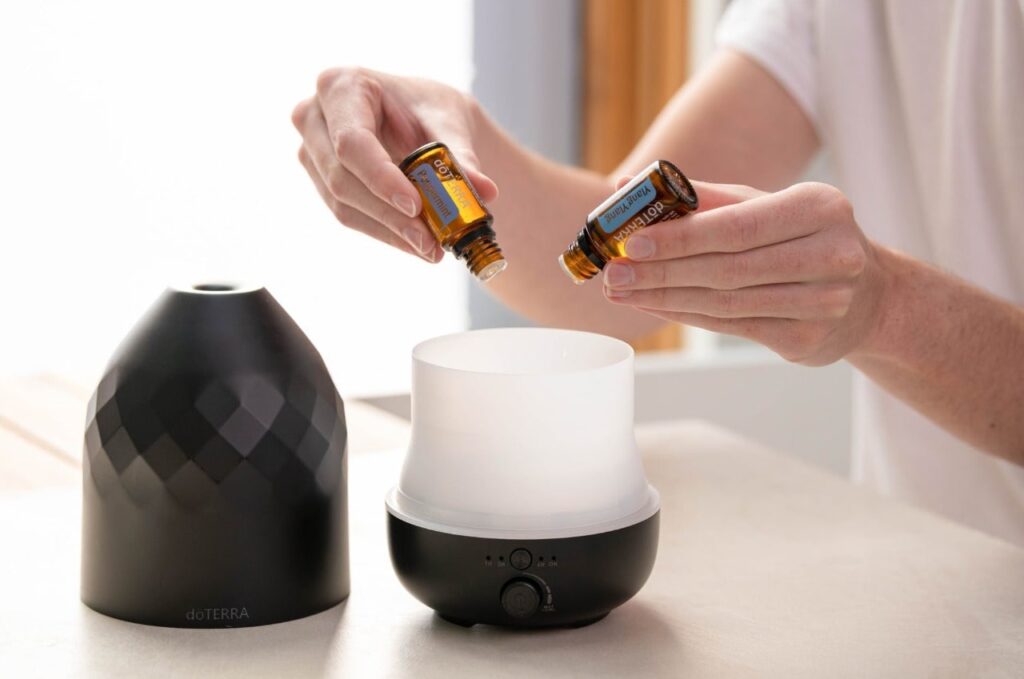A imagem apresenta uma pessoa aplicando dois óleos essenciais em um difusor de cor preta para aplicação da aromaterapia.