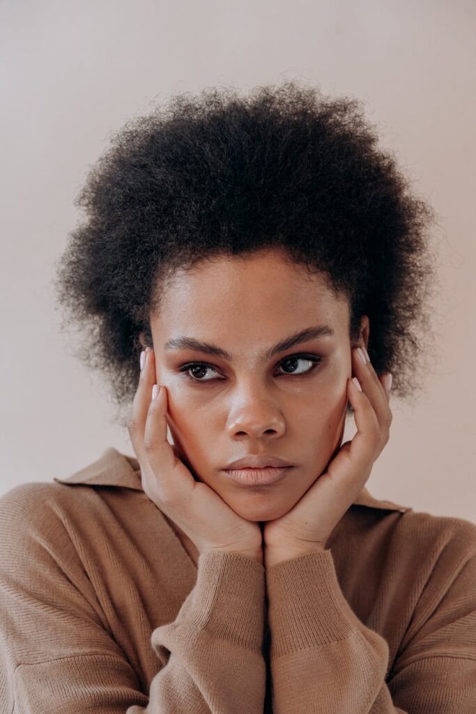 Mulher negra, com as mãos no rosto, olhando para o lado e com cabelo crespo.