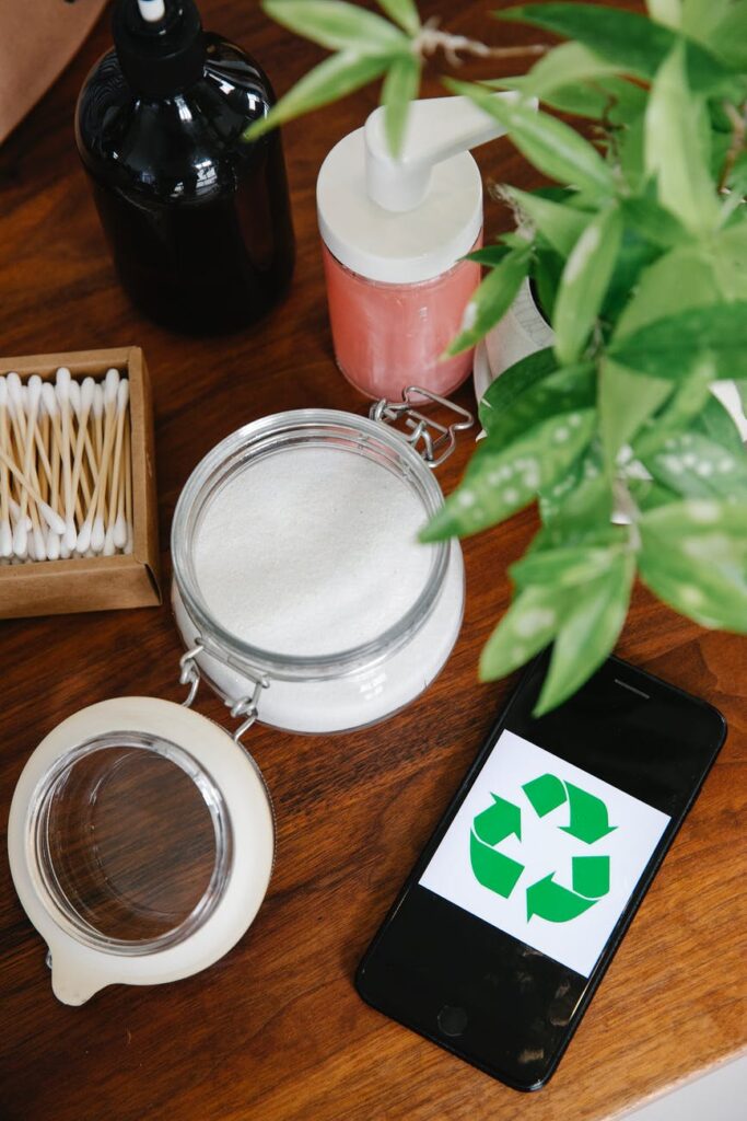 Uma mesa contendo um creme branco dentro de um pote de vidro, algumas embalagens de cosméticos e um celular com o símbolo da reciclagem.